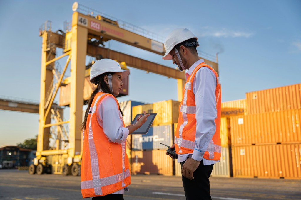 Two workers in hard hats discussing plans near shipping containers